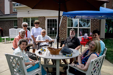 A group of seven elderly and middle-aged people sitting and standing around a round outdoor table under a large umbrella in a courtyard area with brick buildings and windows in the background. There is a blue canopy tent and a table with red tablecloth and decorations behind them.