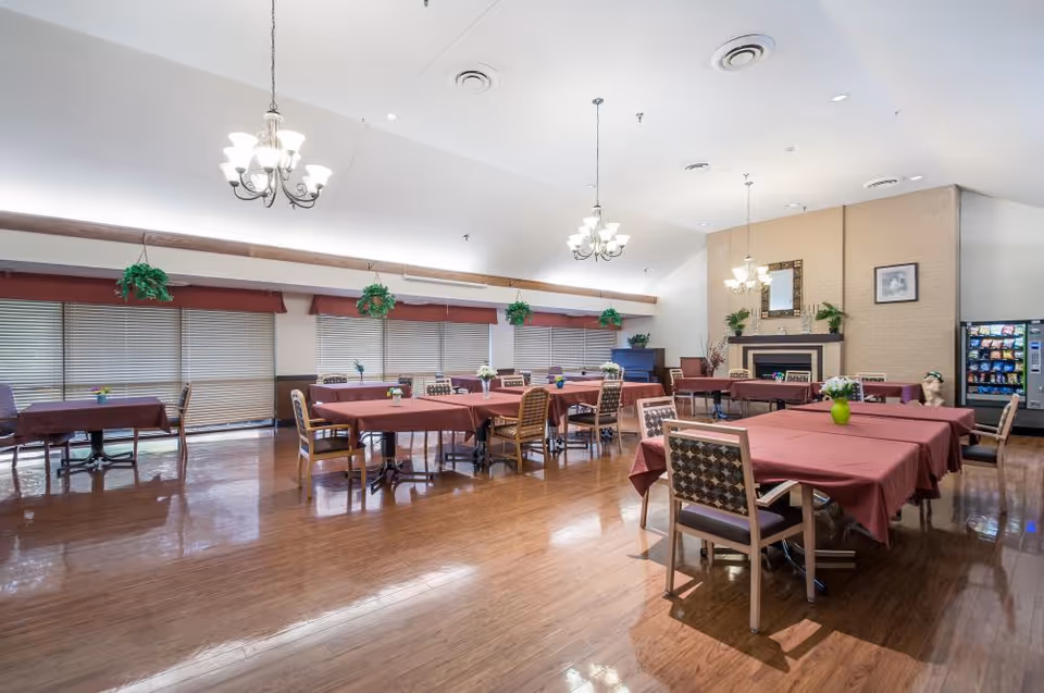 Large communal dining room with tables draped in red cloths, chairs, chandeliers, hanging plants, and a fireplace.