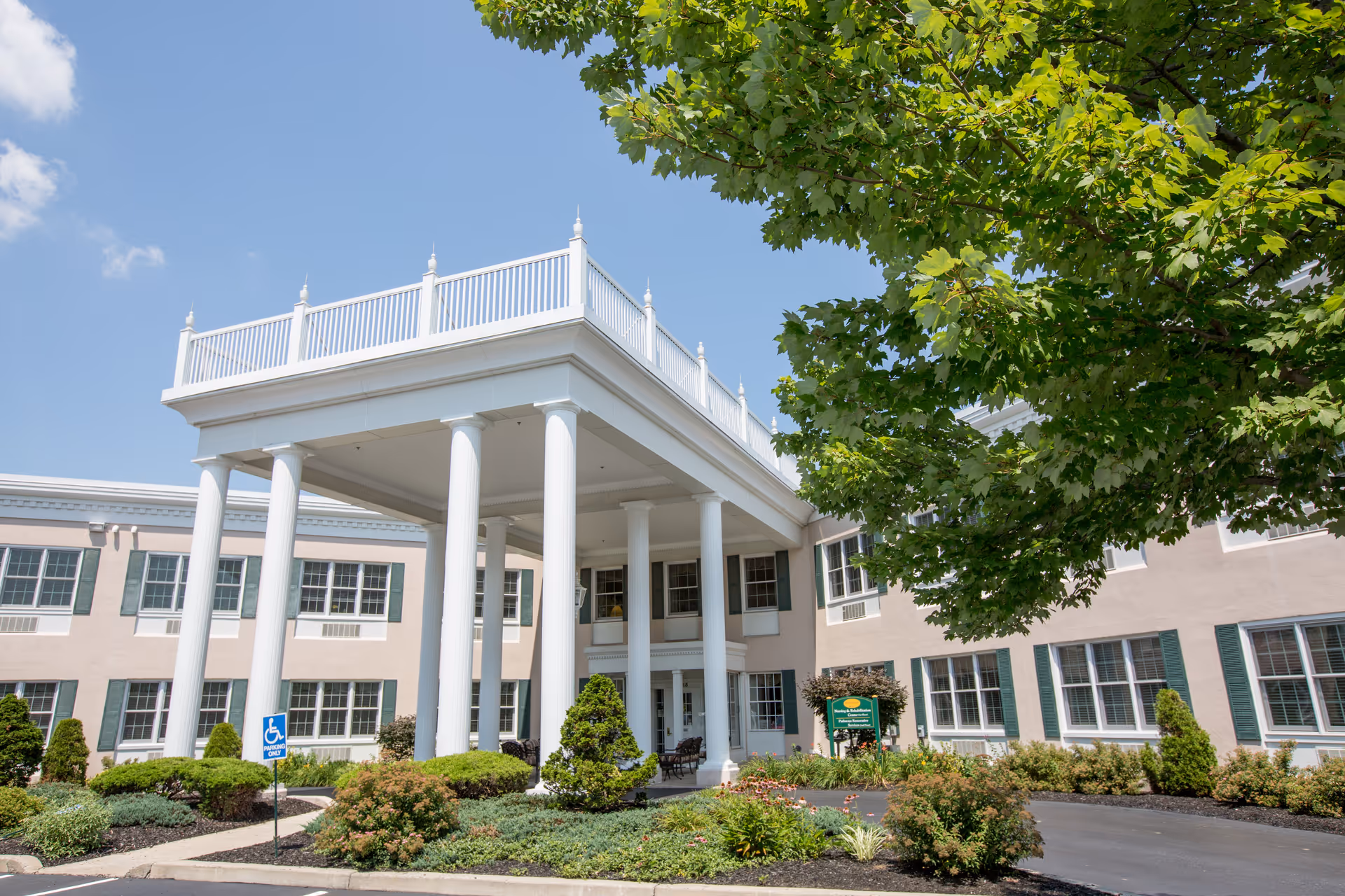Exterior view of a senior living facility building with a large white portico supported by tall columns, surrounded by well-maintained landscaping including bushes and a tree with green leaves under a clear blue sky.