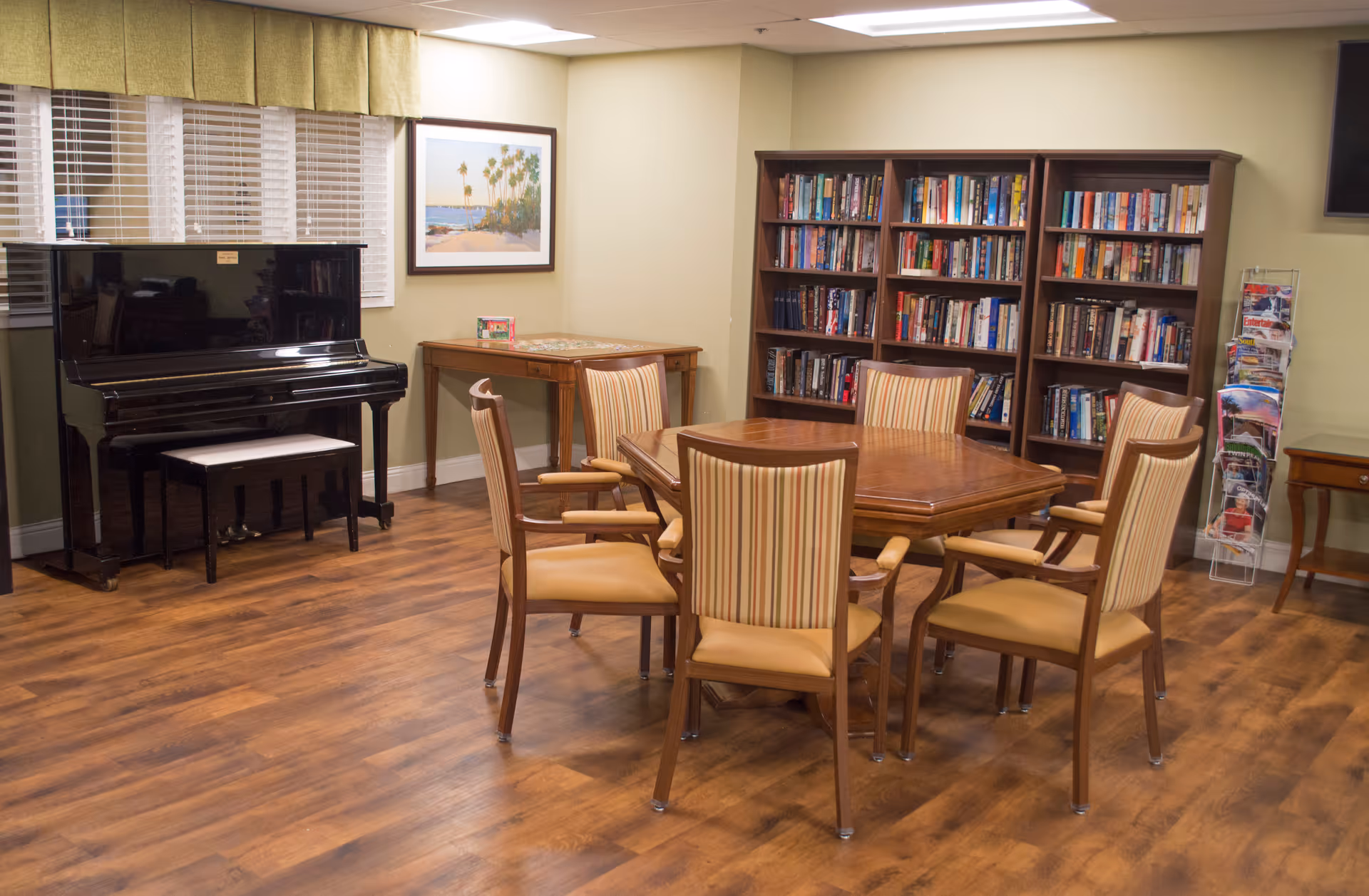 A cozy room with a wooden table surrounded by six striped cushioned chairs, a black upright piano with a bench, a bookshelf filled with books, a small table with a puzzle on it, a magazine rack, and a framed picture of a beach scene on the wall.