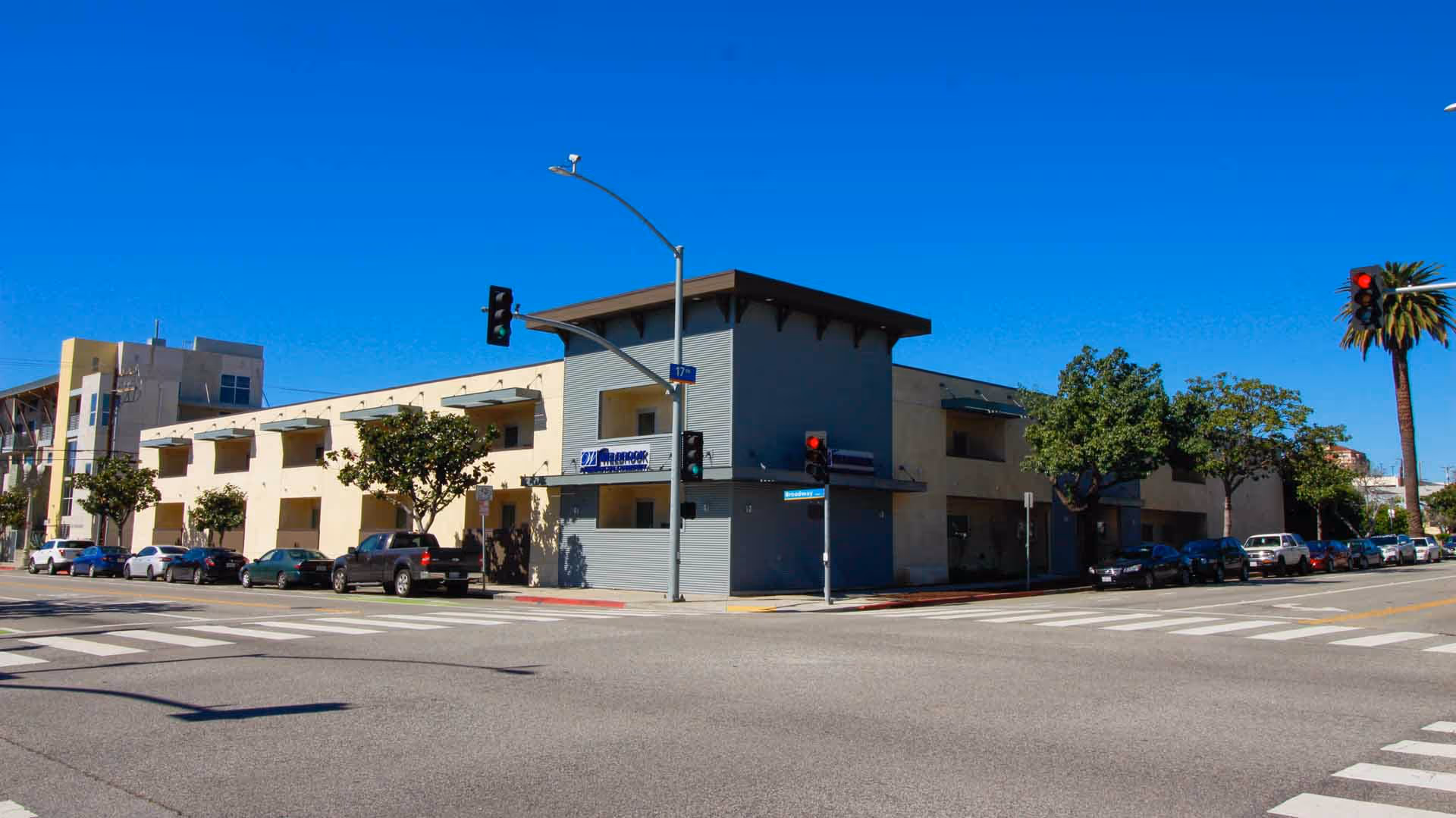 Exterior view of a two-story building on a street corner with parked cars along the sides and traffic lights at the intersection under a clear blue sky.
