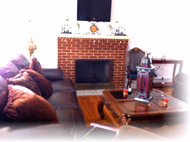 A cozy living room with a brown leather sofa adorned with several brown pillows, a wooden coffee table with decorative items including a lantern and candles, a brick fireplace with a mantel displaying small decorative pieces, and a flat-screen TV mounted above the fireplace. There is also a wooden chair and a small table in the background.
