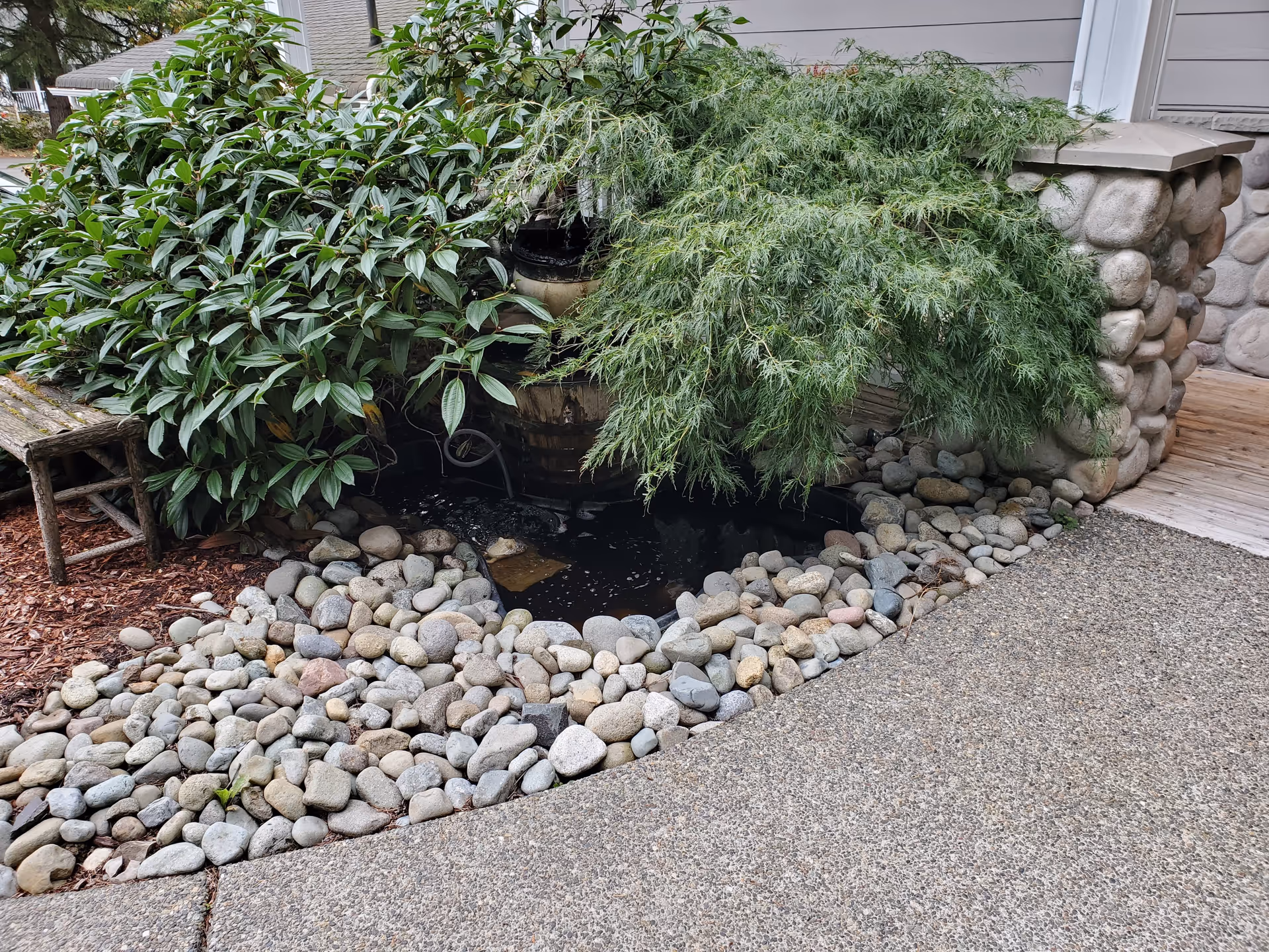 A small outdoor garden area with a water feature surrounded by smooth river rocks, lush green plants, and a stone pillar next to a wooden deck.