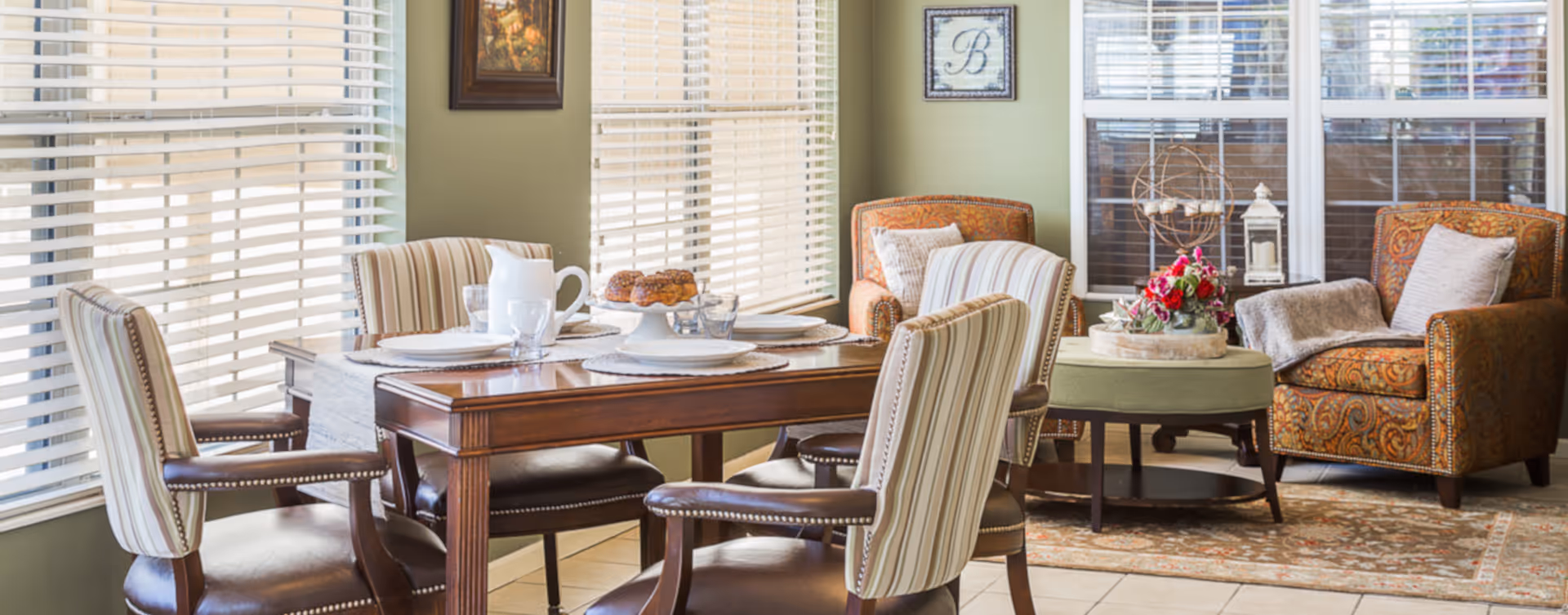 A cozy dining and sitting area with a wooden dining table set for four, featuring striped upholstered chairs. In the background, there are two patterned armchairs with pillows, a round ottoman with a floral arrangement, and large windows with blinds letting in natural light.