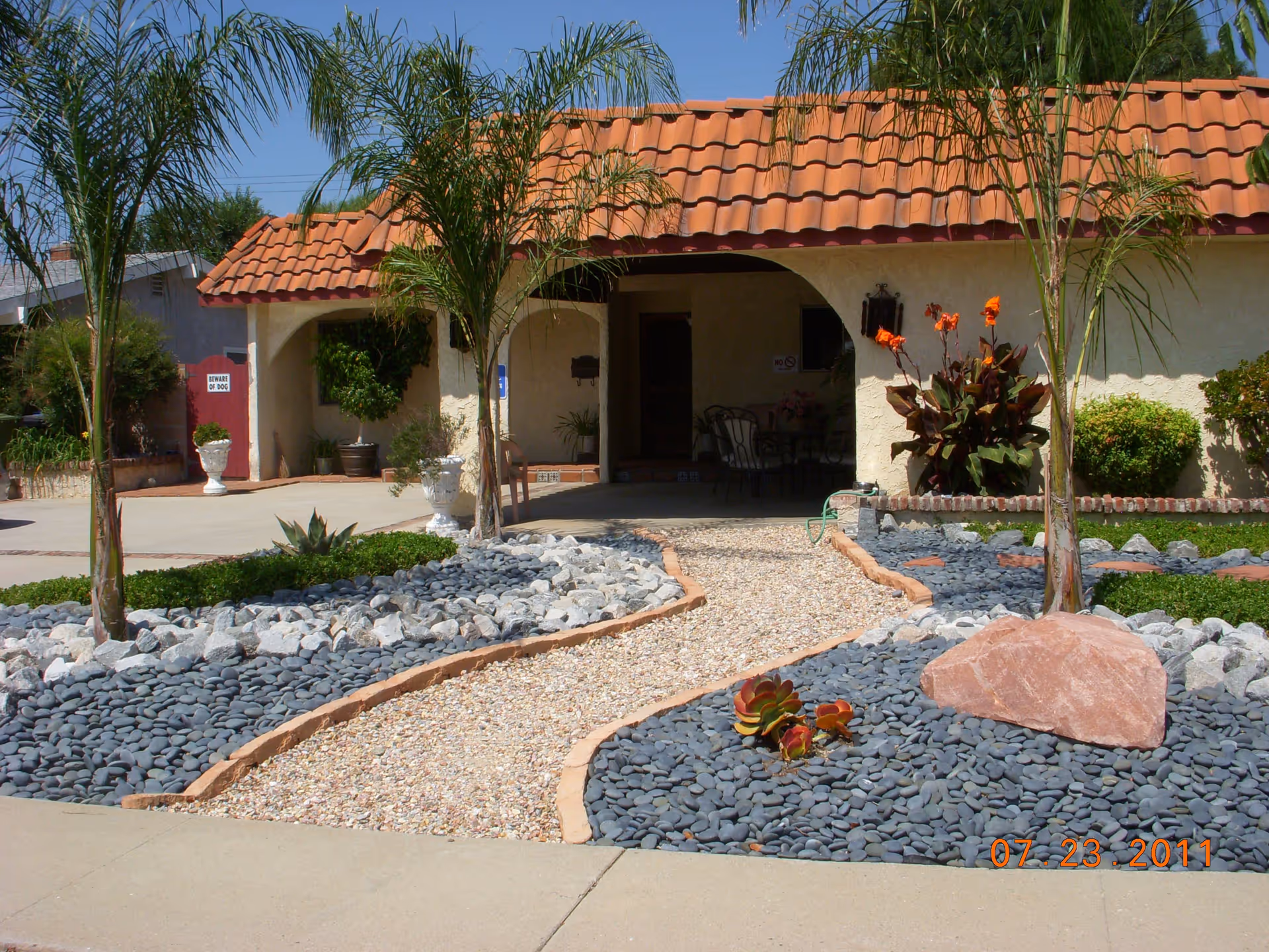 Front exterior view of a single-story building with a red tile roof, a gravel pathway lined with rocks and small palm trees leading to a covered entrance. There are decorative plants and bushes around the pathway and building.