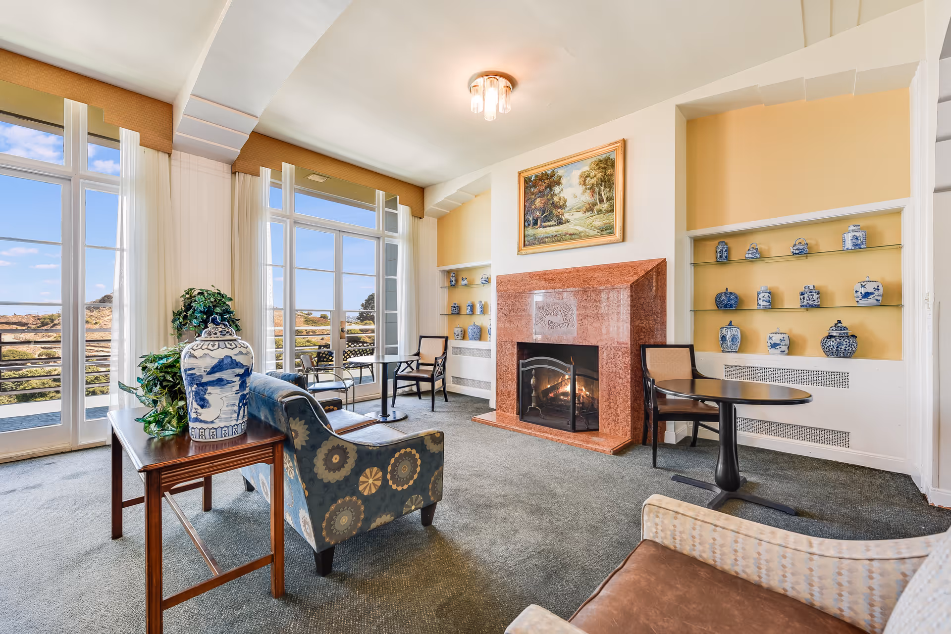 A cozy living room area with large windows showing an outdoor view. The room features a red stone fireplace with a fire burning, a painting above it, and built-in shelves displaying blue and white ceramic vases. There are several chairs and small tables arranged around the room, with a patterned armchair and a wooden side table holding a large blue and white vase and a green plant.