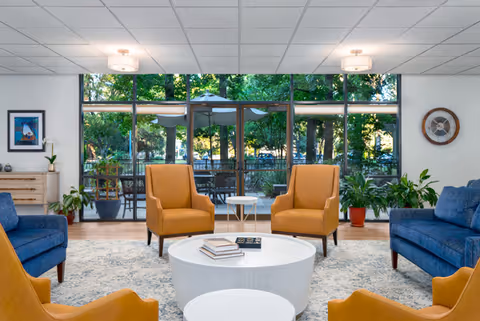 Sunny communal lounge with mustard armchairs and blue sofas arranged around round white coffee tables in front of large floor-to-ceiling windows showing an outdoor patio.