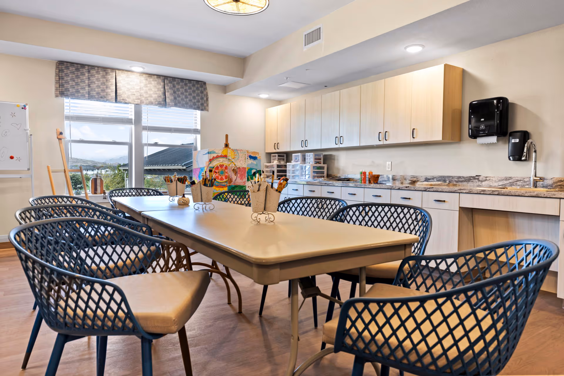 A bright and spacious activity room with a large rectangular table surrounded by eight blue lattice-back chairs with beige cushions. The room features light wood cabinetry along one wall with a granite countertop, a sink, and wall-mounted soap and paper towel dispensers. Large windows with valances let in natural light and offer a view of the outdoors. Art supplies and colorful artwork are visible on the table and in the background.
