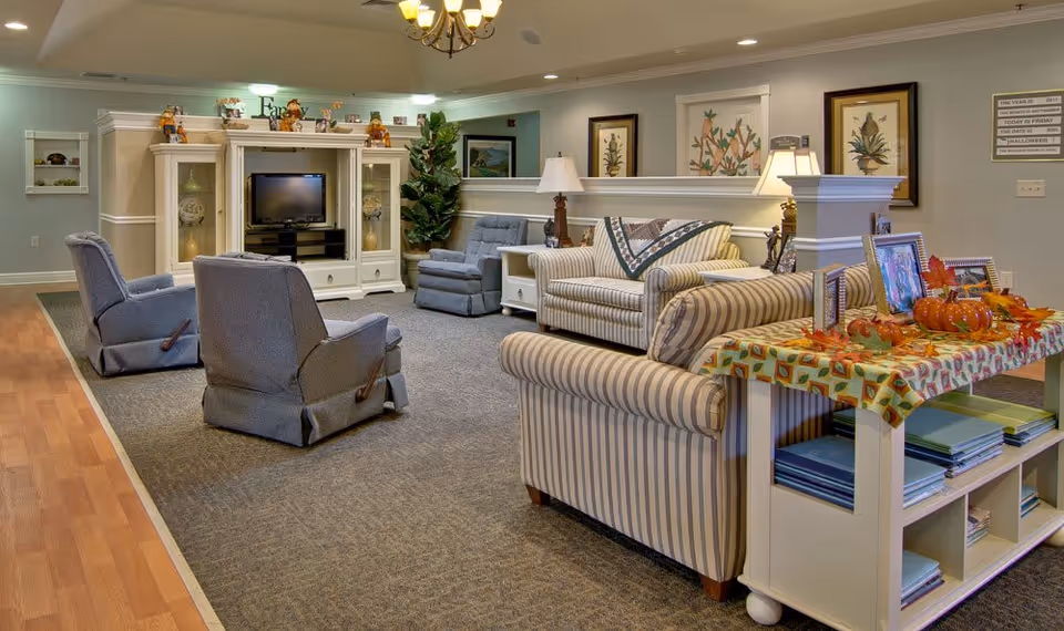 A cozy senior living common area with striped sofas, two blue recliners, a white entertainment center with a TV, decorative plants, framed botanical artwork on the walls, and a table adorned with autumn-themed decorations including small pumpkins and picture frames.