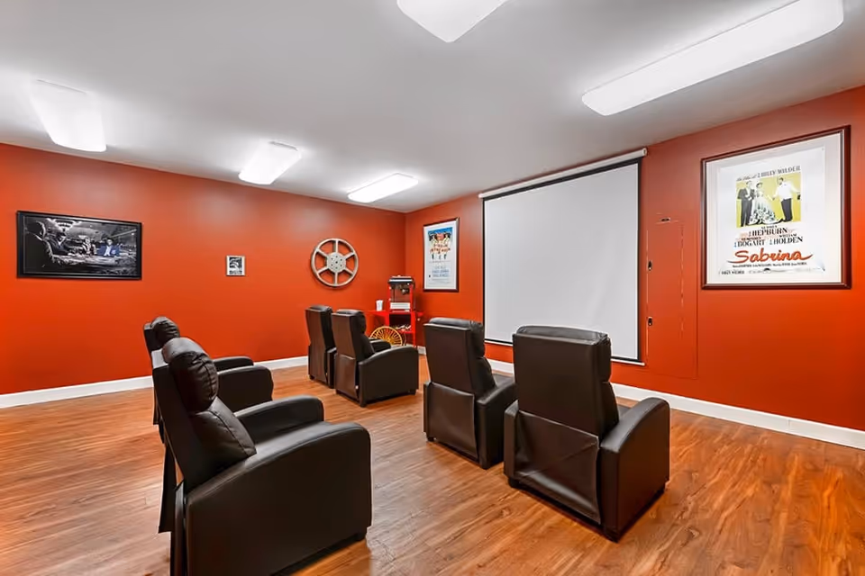 A small movie theater room with six black leather recliners arranged in two rows facing a large white projection screen. The walls are painted red and decorated with framed movie posters and a large film reel. The floor is wood, and the room is lit by ceiling lights.