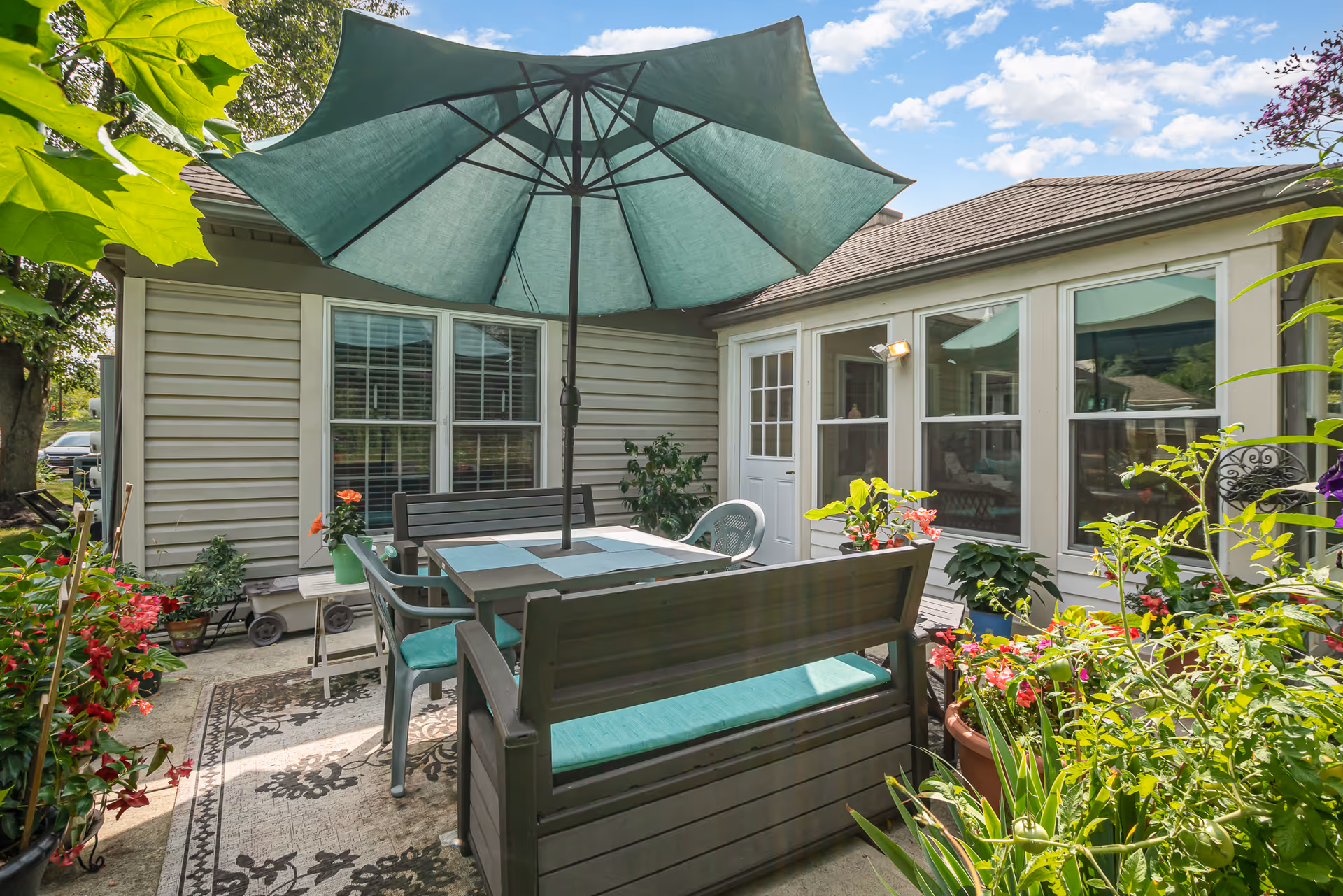Outdoor patio area with a large green umbrella shading a table surrounded by benches and chairs. The patio is decorated with various potted plants and flowers, and the exterior of a building with windows and a door is visible in the background under a partly cloudy sky.
