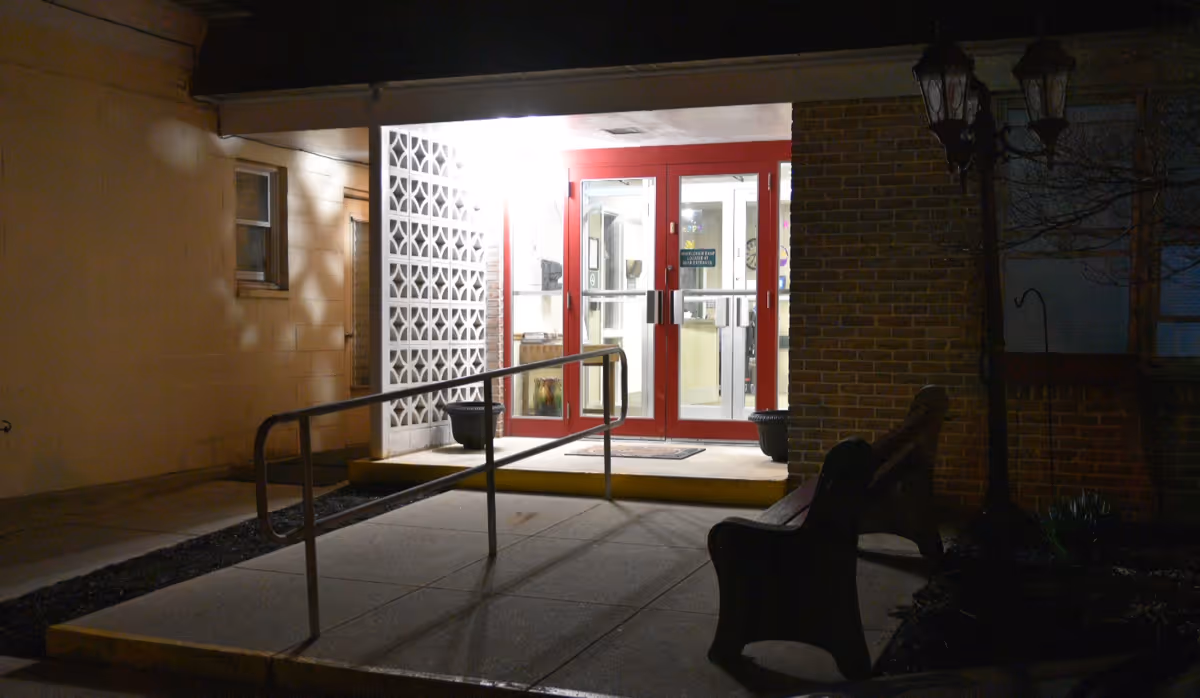 Lit front entrance with red double doors, a wheelchair ramp, and benches outside at night.