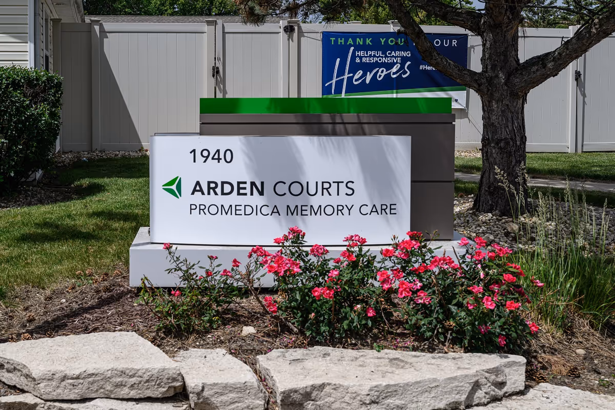 Outdoor view of a sign for Arden Courts ProMedica Memory Care with the address number 1940. The sign is surrounded by pink flowers and landscaping stones, with a white fence and a tree in the background. A blue banner behind the sign reads 'Thank you our helpful, caring & responsive heroes.'
