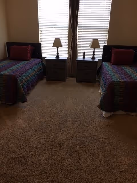 Carpeted bedroom with two twin beds separated by two nightstands and lamps in front of a large window with blinds.