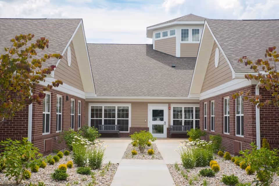 Courtyard entrance of a single-story brick and siding senior living building with a central door, benches, and a landscaped walkway.