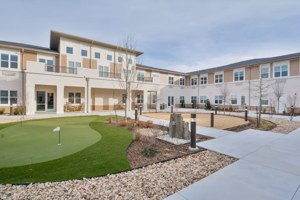 Outdoor courtyard area of Prairie House Assisted Living and Memory Care featuring a small putting green, landscaped garden beds with rocks and shrubs, young trees, and a multi-story building with numerous windows surrounding the courtyard.