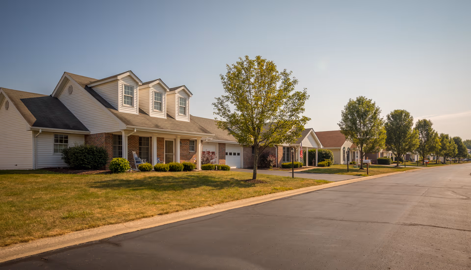 Row of single-story residential units with porches, garages, lawns and trees along a paved street.