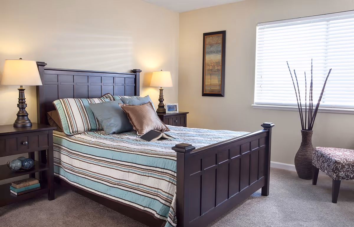 A neatly made bed with a dark wooden frame and striped bedding in shades of blue, brown, and white. There are two matching bedside tables with lamps on each side of the bed. A framed picture hangs on the beige wall, and a window with white blinds allows natural light into the room. A decorative vase with tall sticks and a patterned chair are placed near the window.