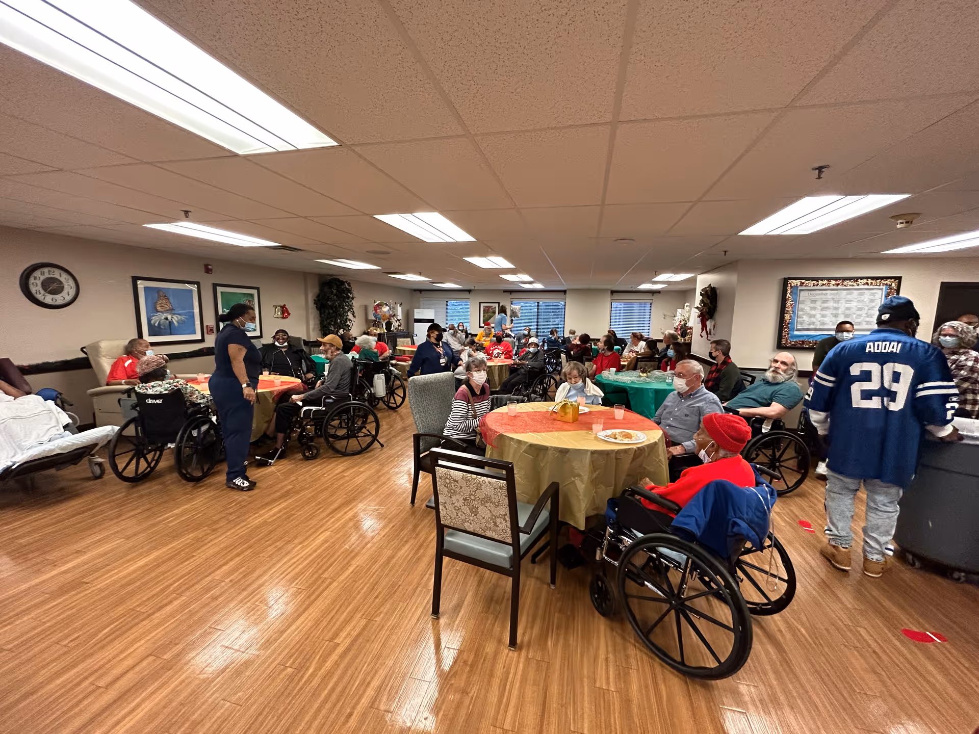 Residents and staff gather around tables in a large communal dining/activity room, many seated in wheelchairs.