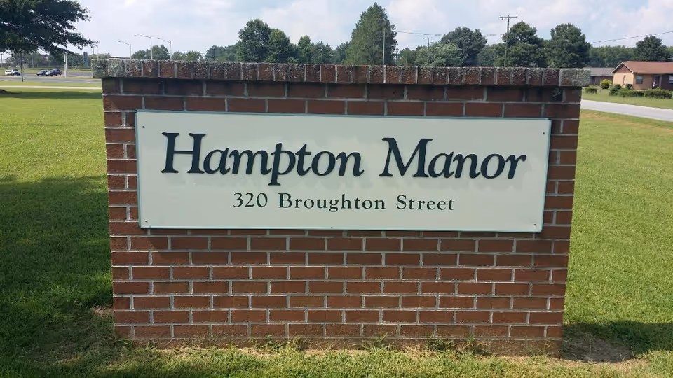 Brick entrance sign reading "Hampton Manor 320 Broughton Street" on a grassy lawn.