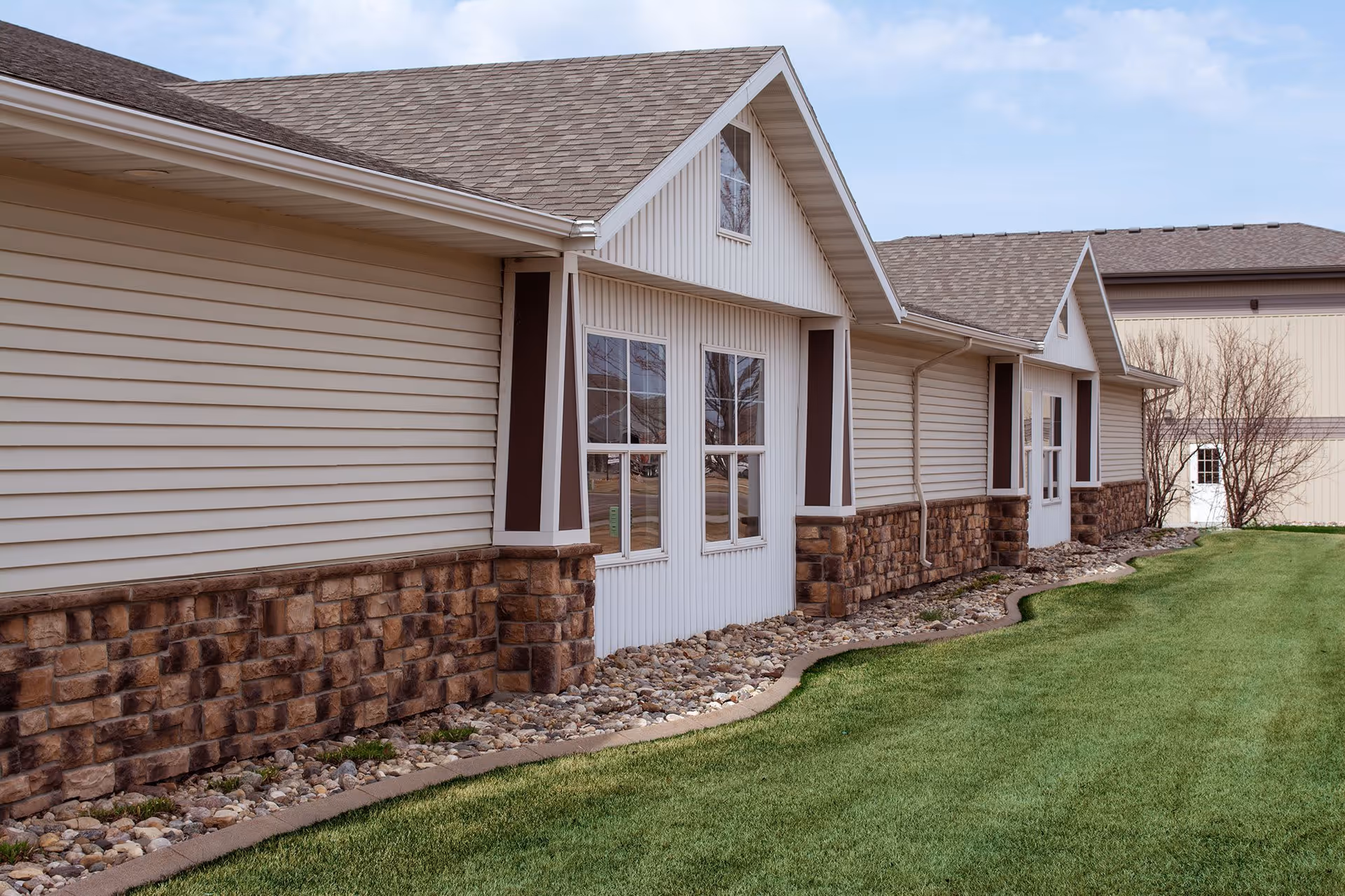 Exterior view of a single-story building with beige siding and stone accents along the lower walls. The building has multiple windows and a well-maintained green lawn with a rock border along the foundation. The sky is partly cloudy.