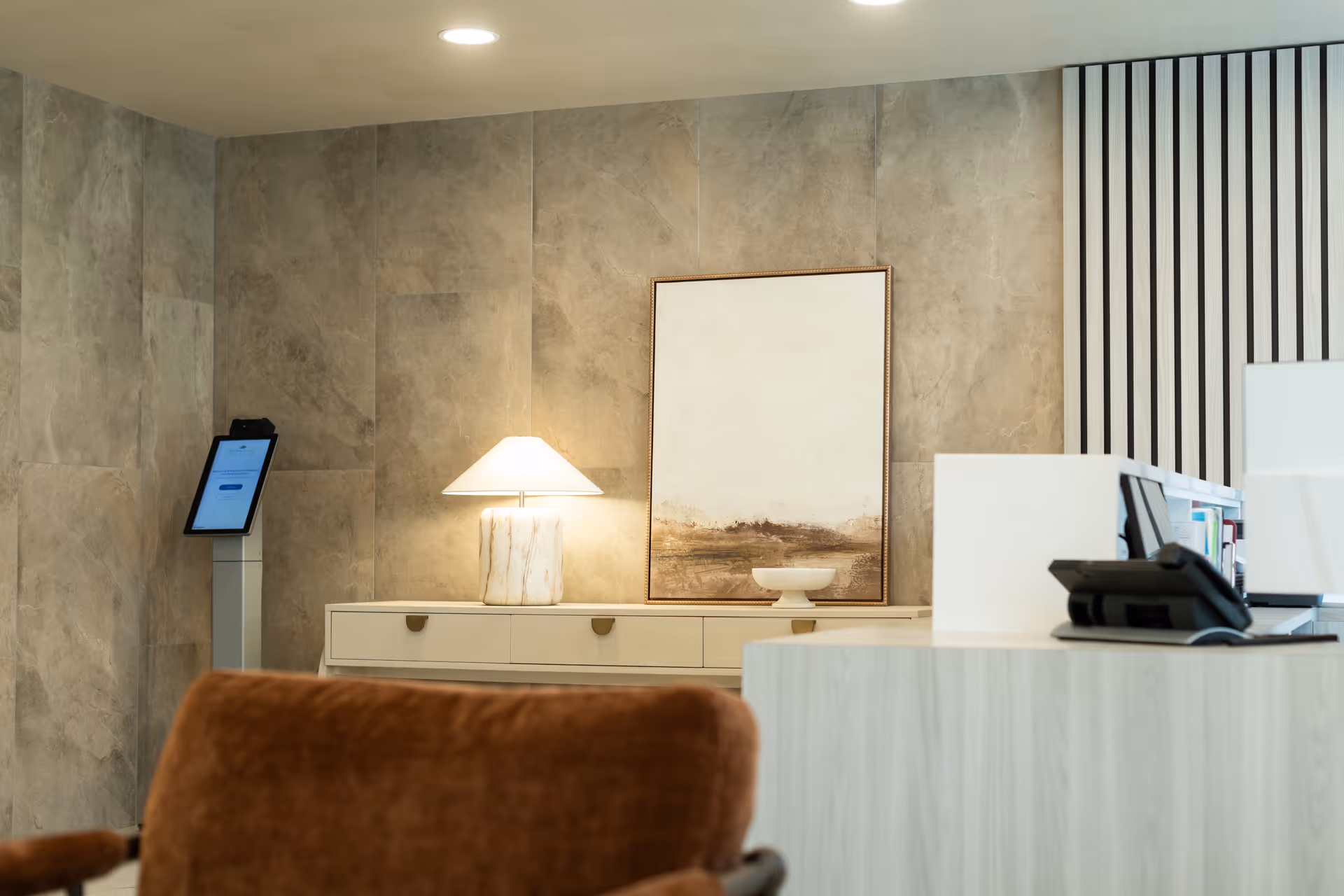 A modern reception area with a light-colored wooden desk, a brown upholstered chair in the foreground, a table lamp with a white shade on a side cabinet, and a large abstract painting on the wall behind. The walls have a stone-like texture, and there is a digital kiosk or tablet stand to the left.