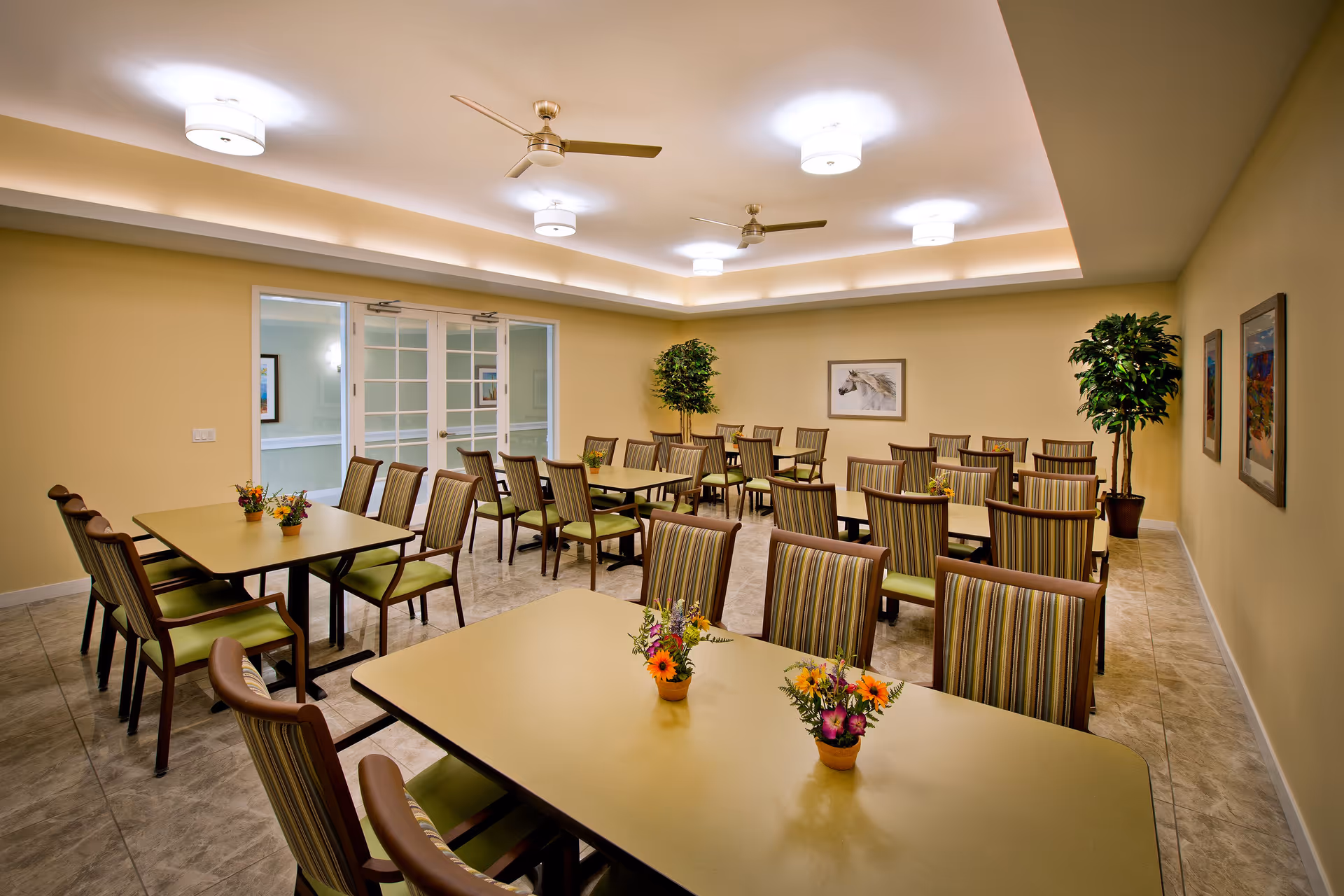 A well-lit dining room with multiple tables and chairs arranged neatly. Each table has small flower arrangements as centerpieces. The room features beige walls, a tiled floor, ceiling fans, and modern ceiling lights. There are framed pictures on the walls and potted plants in the corners. Double glass doors are visible at the back of the room.