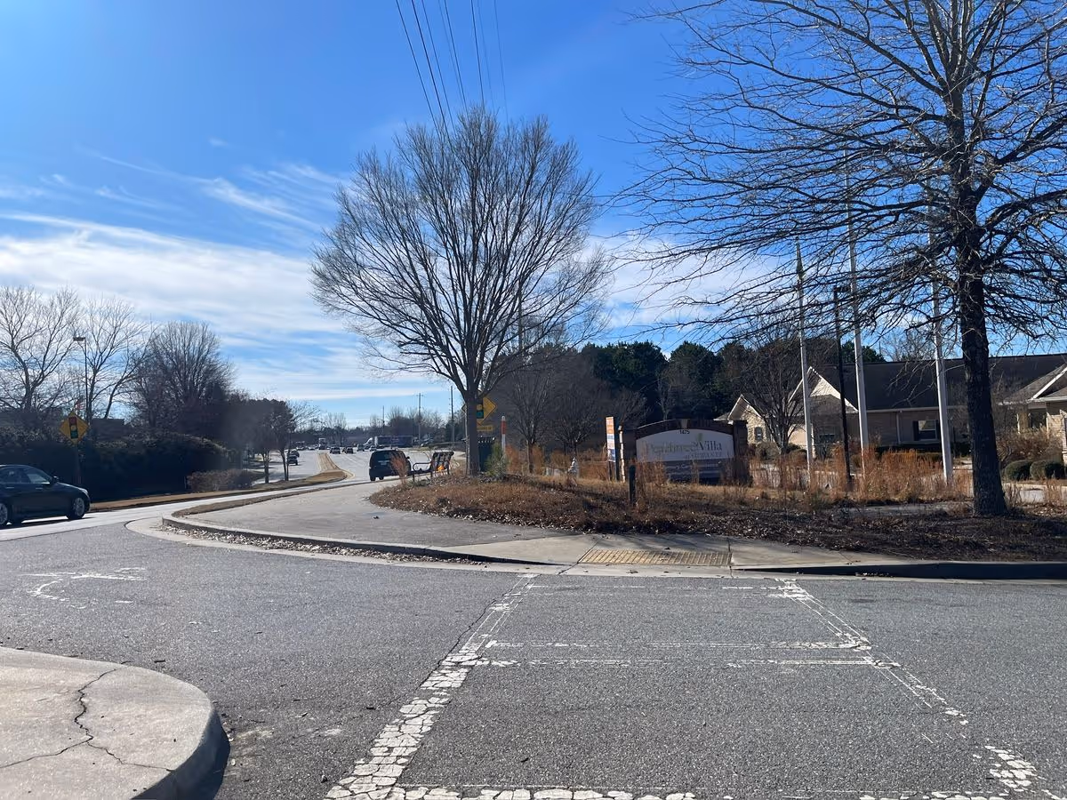 Entrance to Peachtree Villa at Suwanee seen from a road with a landscaped median, trees, and a few cars under a clear blue sky.