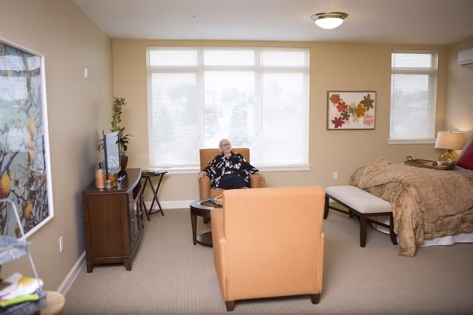 A cozy bedroom with beige walls and carpet, featuring a bed with a brown comforter and a bench at the foot. There are two orange armchairs facing each other with a small round table between them. A woman with white hair and glasses is sitting in one of the armchairs, smiling. The room has large windows with blinds, a wooden TV stand with a television, a lamp on a side table, and a framed floral artwork on the wall.