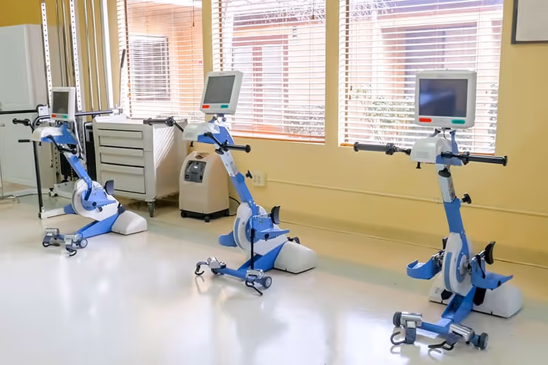 Three blue and white stationary exercise bikes with digital screens are positioned in a bright room with large windows covered by wooden blinds. The floor is shiny and light-colored, and there is a white cabinet and a small medical device against the wall.