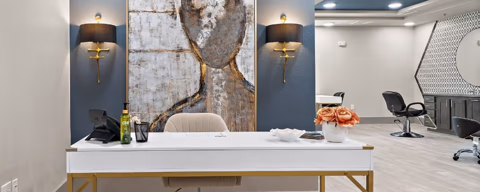 A modern reception or front desk area with a white desk featuring gold accents, a beige chair, decorative items including a vase with orange flowers, a bottle, and a small bowl. Behind the desk is a large abstract painting and two wall sconces with black shades. In the background, there are black salon chairs and a large round mirror on a patterned wall, suggesting a salon or beauty area within the facility.