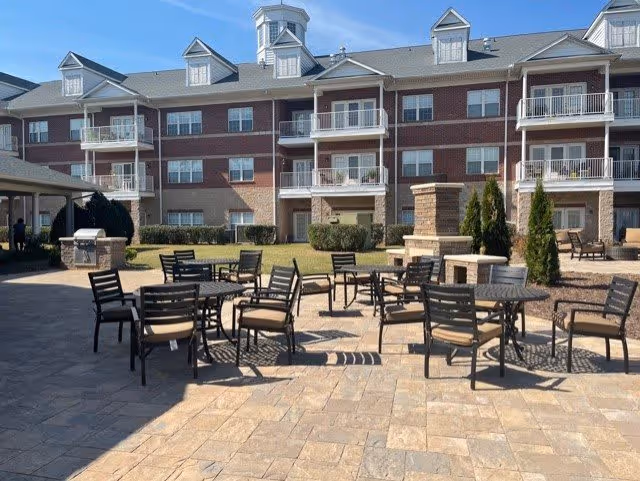 Outdoor patio area with multiple black metal tables and chairs with beige cushions, set on a stone-paved surface. In the background is a three-story brick and stone building with balconies and dormer windows under a clear blue sky.