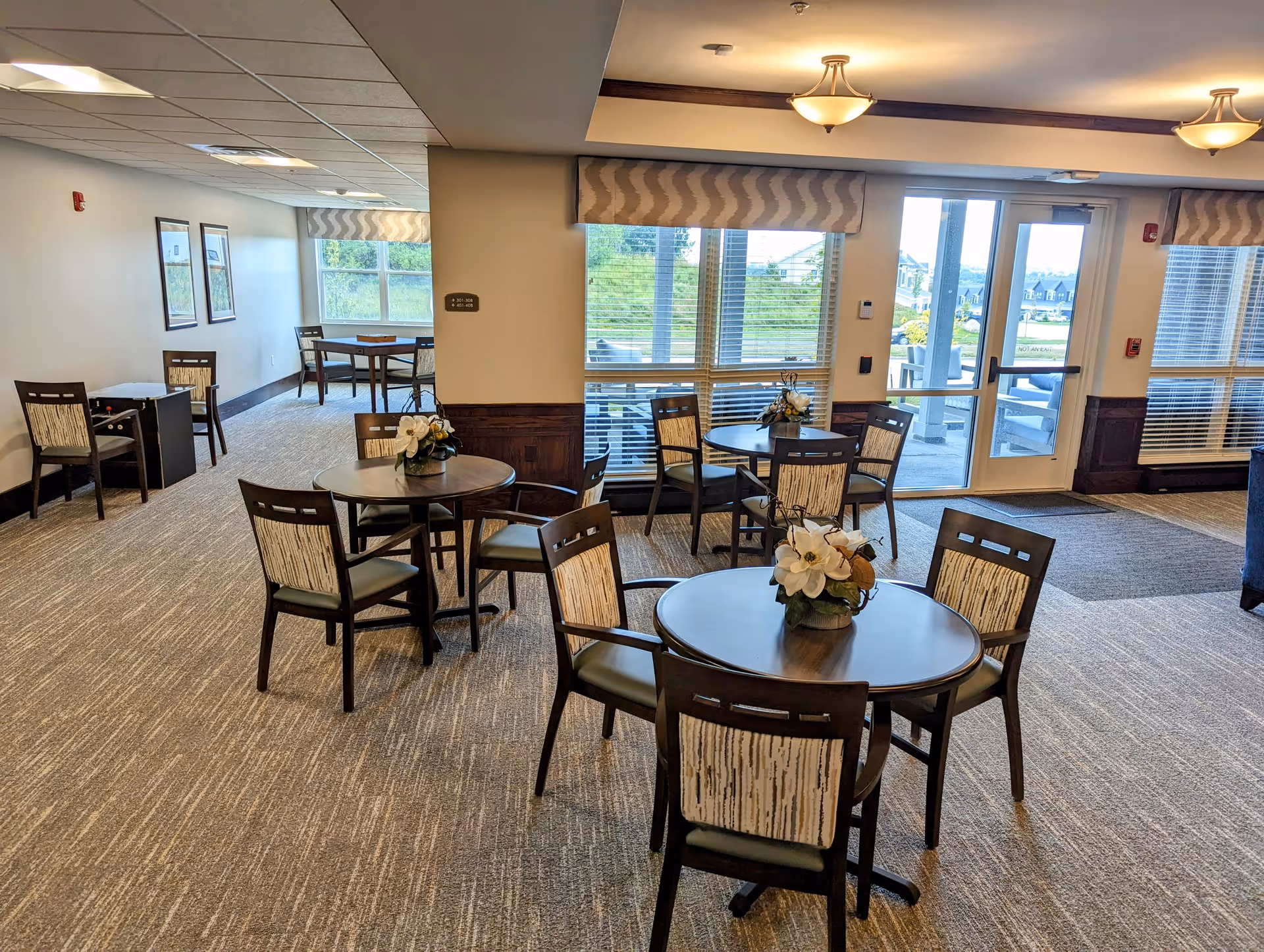 A bright and spacious dining area with several round wooden tables, each surrounded by four chairs with patterned backs and cushioned seats. Each table has a small floral centerpiece. Large windows and glass doors allow natural light to fill the room and provide a view of an outdoor patio area with seating. The room has carpeted floors, beige walls, and ceiling lights with warm tones.