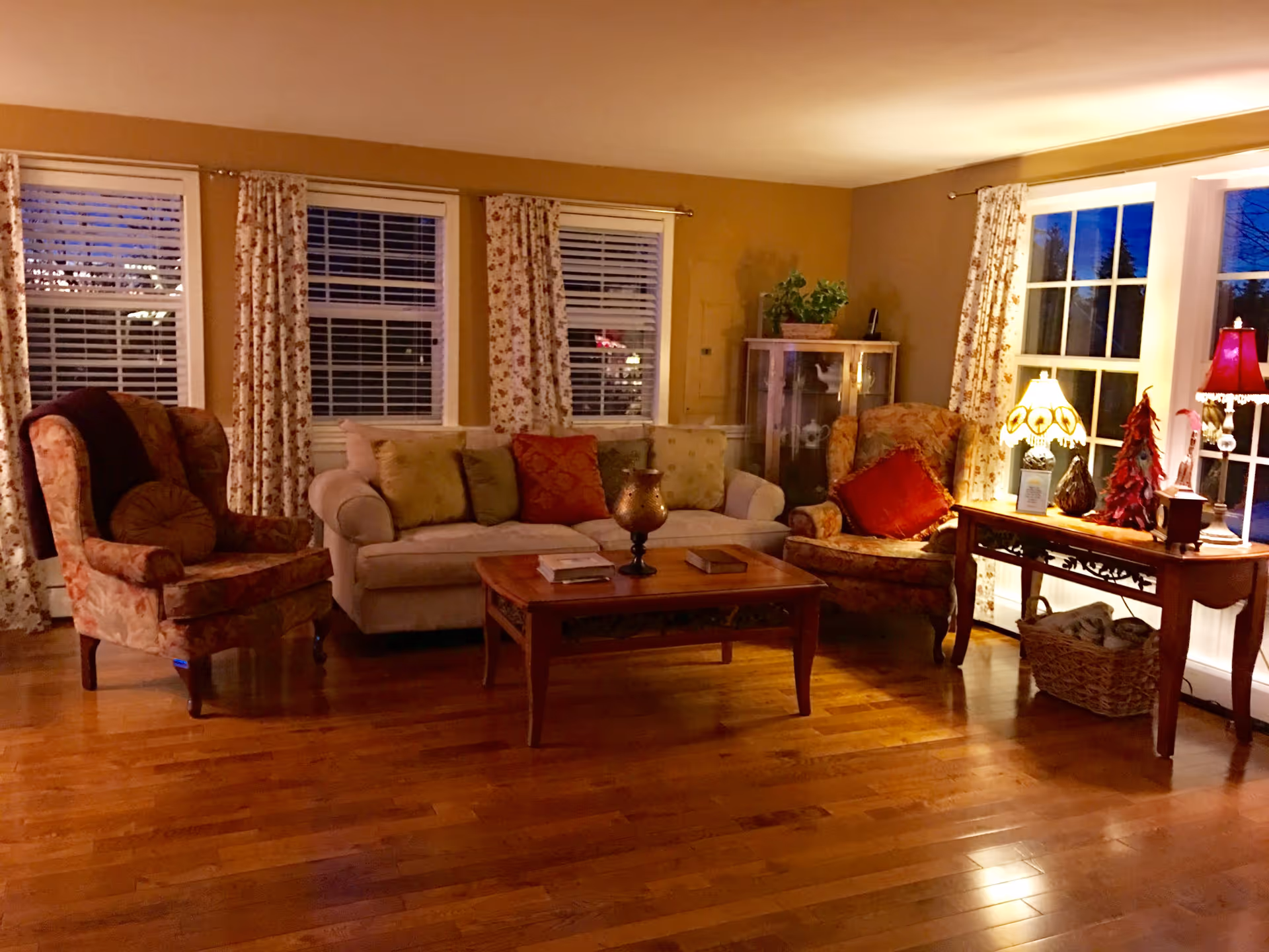 A cozy living room with wooden flooring, a beige sofa with multiple cushions, two floral-patterned armchairs, a wooden coffee table with books and a decorative item, and a wooden side table with two lamps and decorative pieces. The room has large windows with floral curtains and warm lighting.