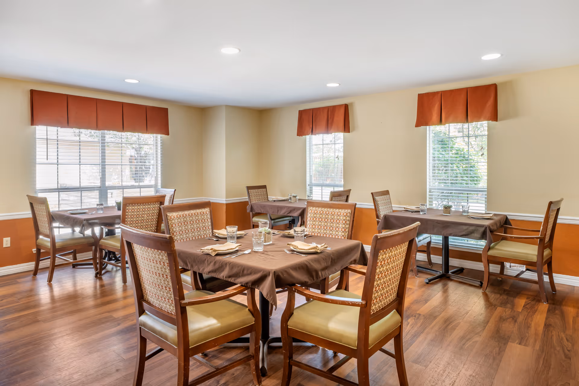 A dining room with several wooden tables covered with brown tablecloths, each set with glasses, napkins, and utensils. The room has wooden flooring, beige walls with an orange lower section, and windows with orange valances letting in natural light.