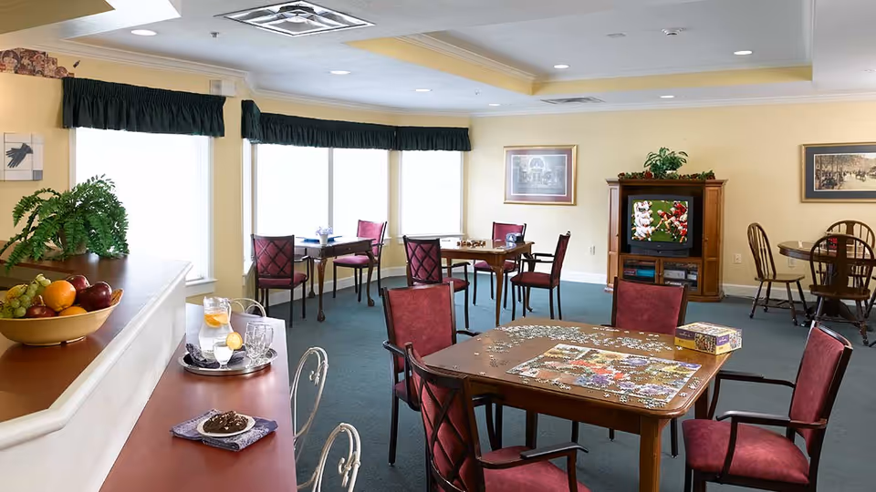 A bright common room with multiple tables and chairs, some with puzzles and board games on them. A television is on in the corner, and large windows with dark green valances let in natural light. A counter with a bowl of fruit and drinks is visible in the foreground.