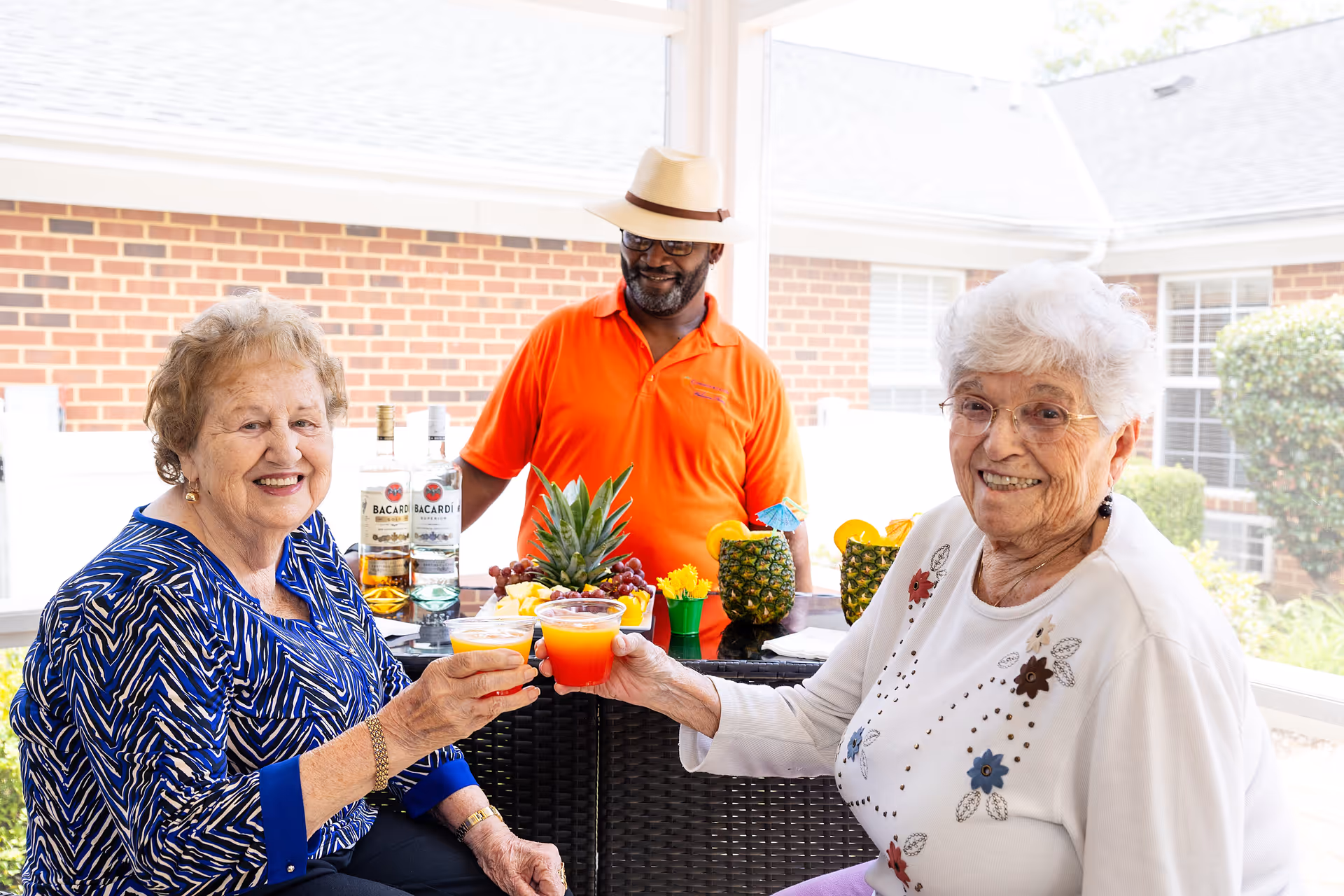 Two elderly women sitting outdoors at a table, smiling and holding colorful drinks in a toast. Behind them, a man wearing an orange shirt and a hat stands near a table with tropical fruits and bottles of Bacardi rum. The setting appears to be a covered patio area with brick walls and windows in the background.