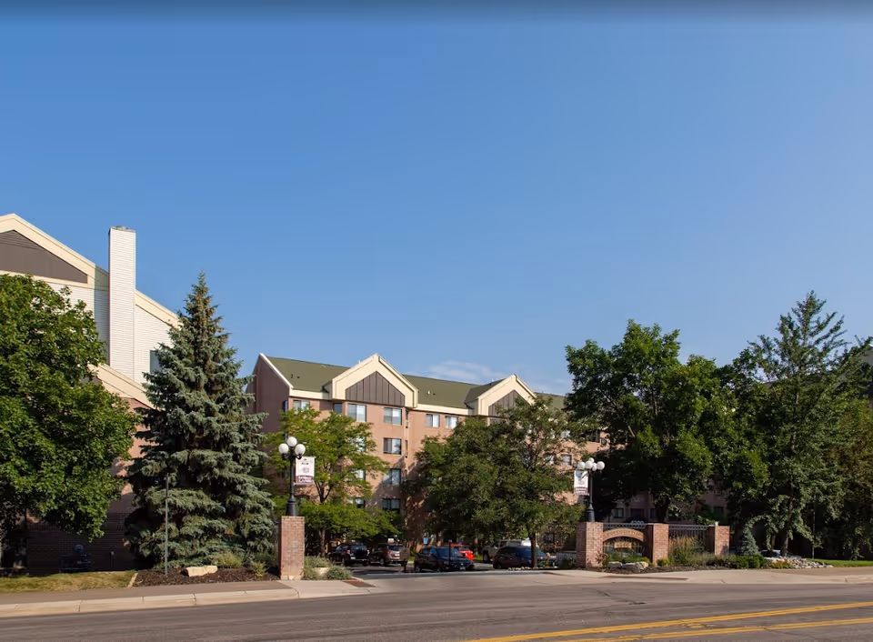Exterior view of a multi-story senior living facility building with a green roof and brick facade, surrounded by trees and a parking area in front under a clear blue sky.