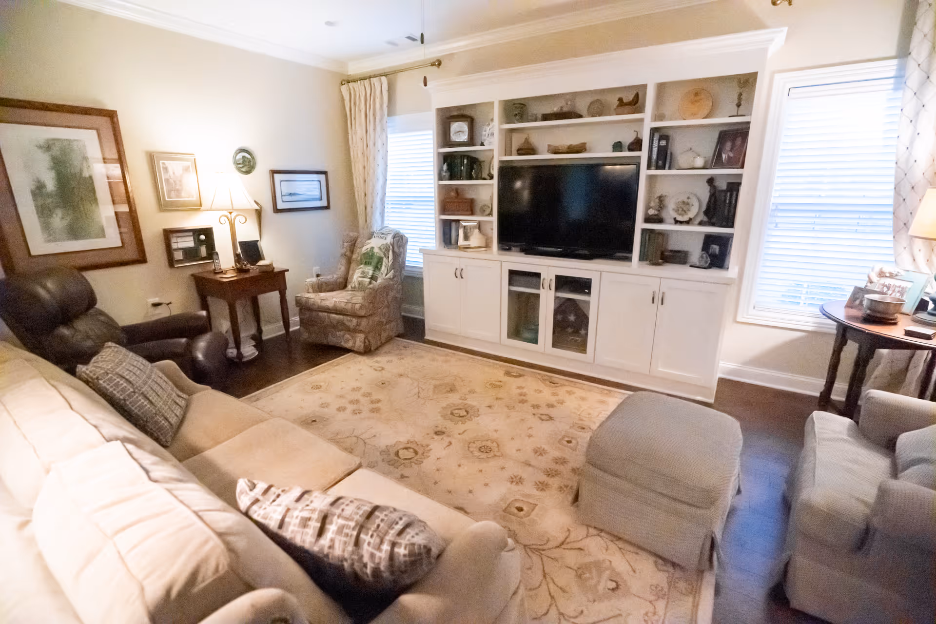 A cozy living room with a beige sofa, a dark brown recliner, a patterned armchair, and a light blue armchair. There is a large built-in white entertainment center with shelves and cabinets, housing a flat-screen TV and various decorative items. The room has two windows with blinds and curtains, a wooden side table with a lamp, and a beige patterned area rug covering the dark hardwood floor.