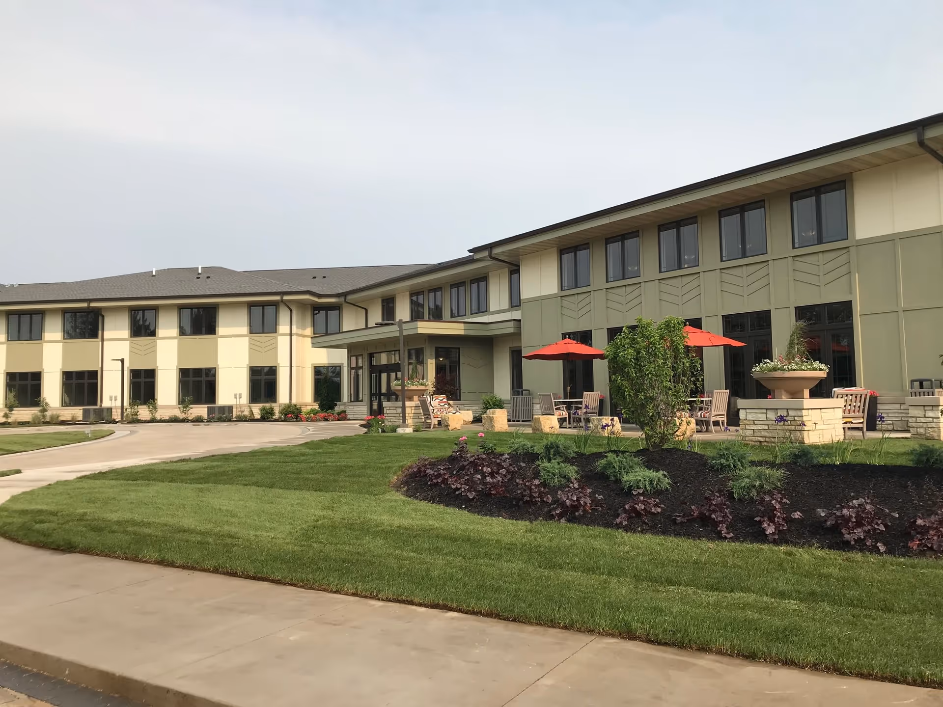 Exterior front view of a two-story senior living building with a landscaped lawn, patio seating and red umbrellas.
