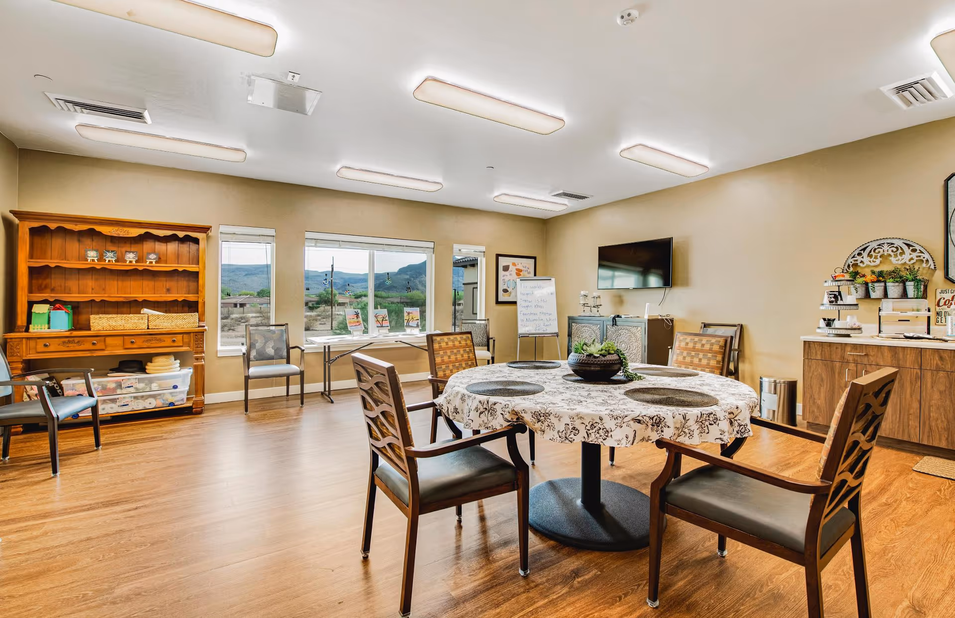 A bright and spacious common room with a round table covered with a floral tablecloth and four chairs around it. The room has wooden flooring and beige walls. There is a wooden hutch with baskets and decorative items on the left side, and a sideboard with plants and coffee supplies on the right. Large windows offer a view of a mountainous landscape outside. A flat-screen TV is mounted on the wall, and a whiteboard with writing is visible near the windows.