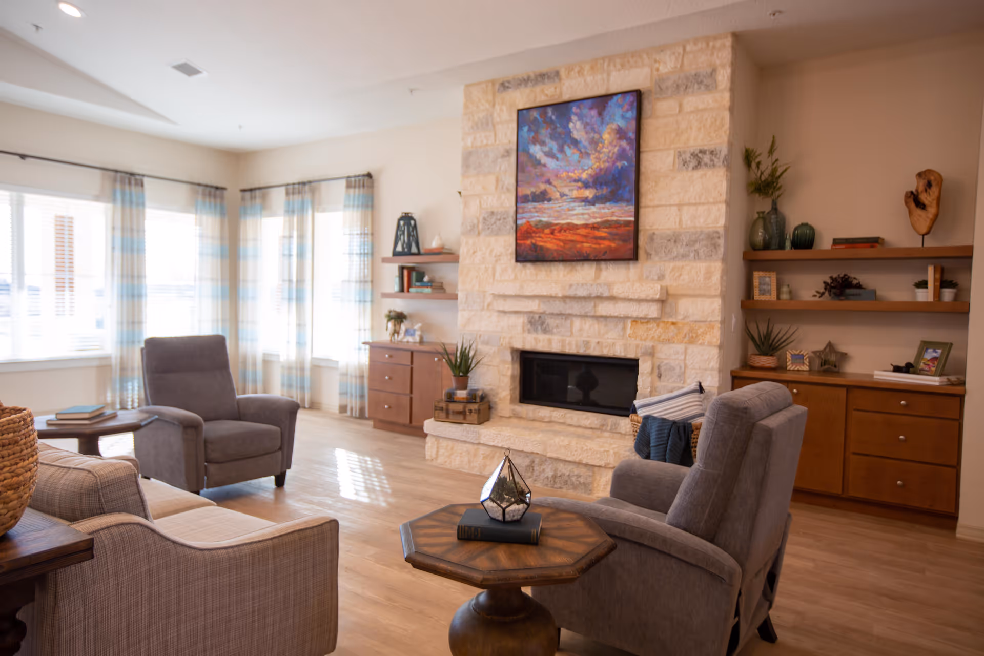 Bright common living room with a stone fireplace, comfortable armchairs, wooden side tables and built-in shelving.