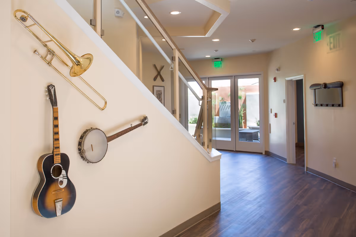 Interior hallway of Vista Rosa Elder Care with musical instruments including a trombone, guitar, and banjo mounted on a beige wall next to a staircase with glass railing. The hallway has wood flooring, recessed ceiling lights, and glass doors leading to an outdoor area with plants. Exit signs are visible above the doors.