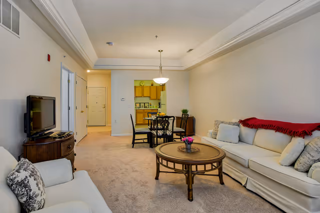 A cozy living room and dining area in a senior living facility. The living room features a white sofa with multiple cushions and a red throw blanket, a round wooden coffee table with a small flower arrangement, and a white armchair with a patterned pillow. The dining area has a round table with four chairs and a hanging light fixture above. The kitchen is visible in the background with wooden cabinets and a refrigerator. The walls are painted light beige, and the floor is carpeted.