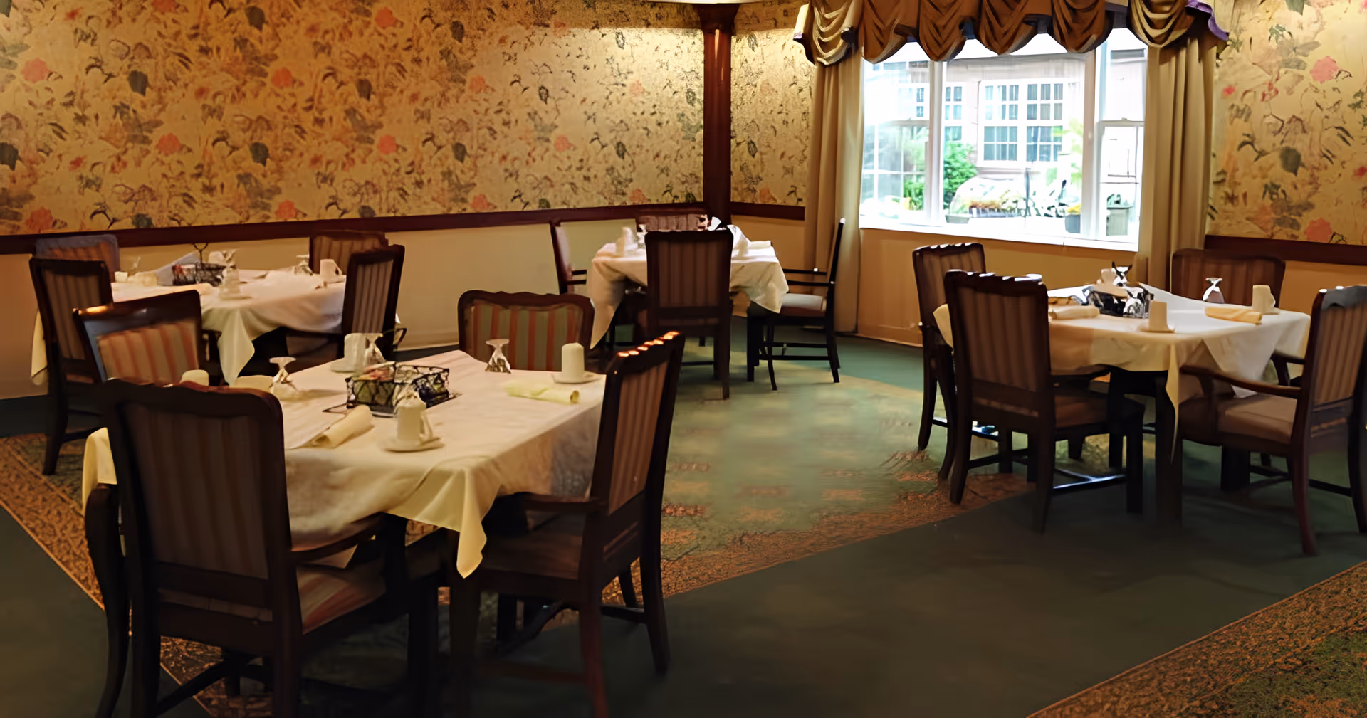 A dining room with several tables covered in white tablecloths, each set with napkins, cups, and glasses. The room has floral wallpaper, green carpet with patterned borders, wooden chairs with striped upholstery, and a large window with curtains letting in natural light.