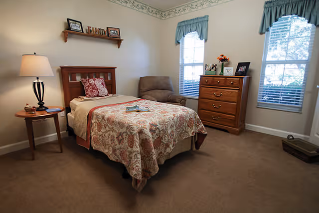 A cozy bedroom featuring a single wooden bed with a patterned bedspread and pillows, a wooden nightstand with a lamp, a comfortable armchair, a wooden dresser with decorative items, and two windows with blue valances letting in natural light.