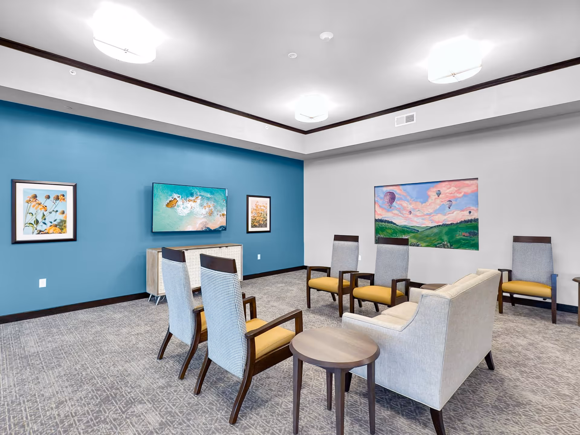 A modern common area with a blue accent wall featuring a mounted flat-screen TV and two framed floral pictures. The room has several chairs with yellow cushions and a beige sofa arranged around small wooden tables. Another wall has a colorful painting of hot air balloons over green hills under a pink sky. The carpet is patterned in neutral tones, and the ceiling has three large, round light fixtures.