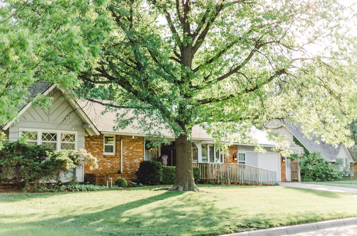 Front of a single-story brick-and-siding house with a large leafy tree and a green lawn.