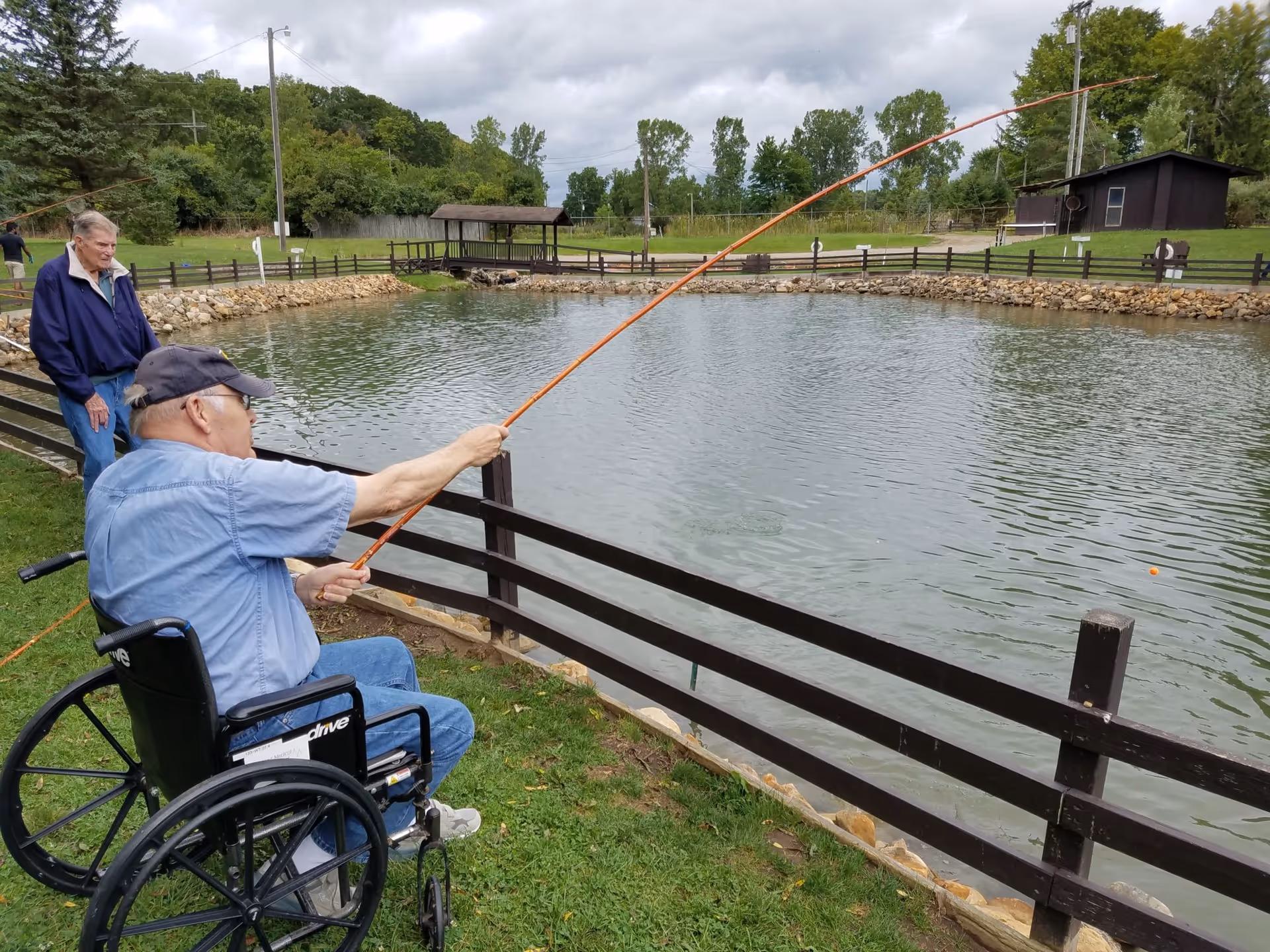 An elderly man in a wheelchair fishing with a fishing rod by a pond, accompanied by another elderly man standing nearby. The scene is outdoors with green grass, trees, and a cloudy sky.