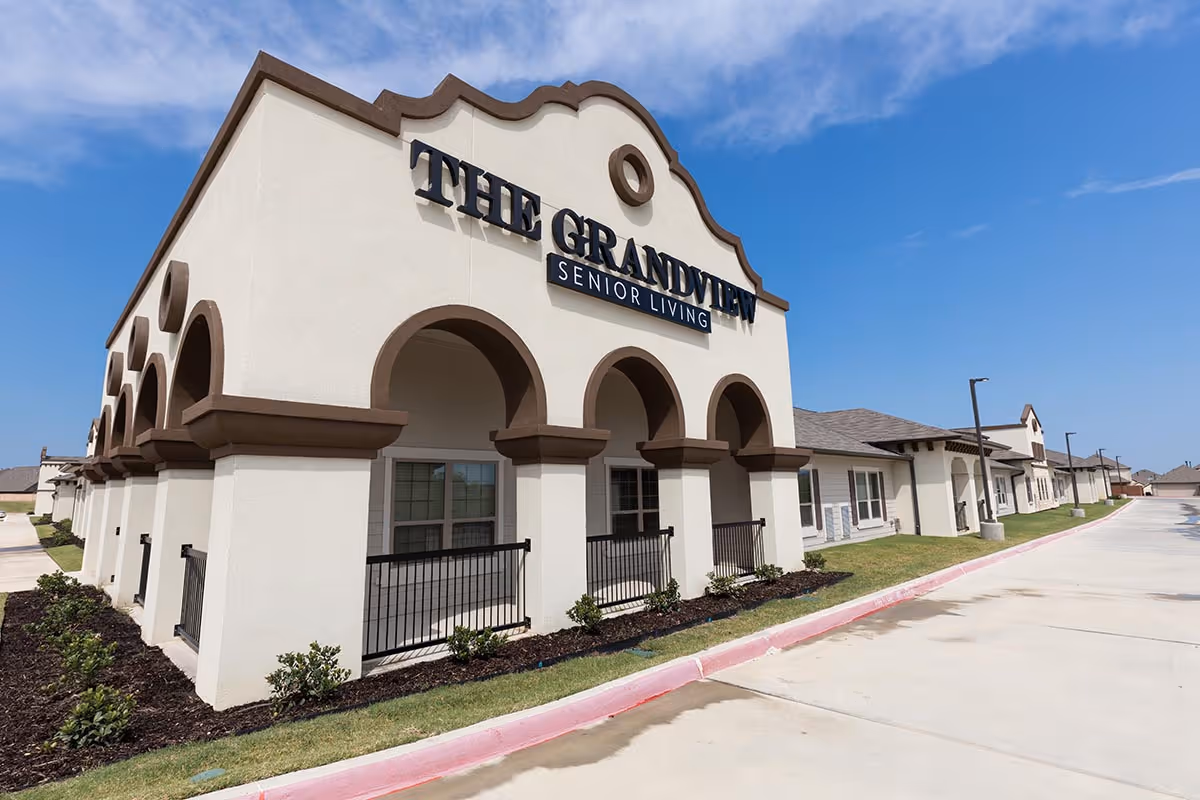 Exterior view of The Grandview Senior Living facility building with arched architectural features, beige walls with brown trim, and a clear blue sky above. The building is surrounded by a paved driveway and landscaped with small bushes and grass.